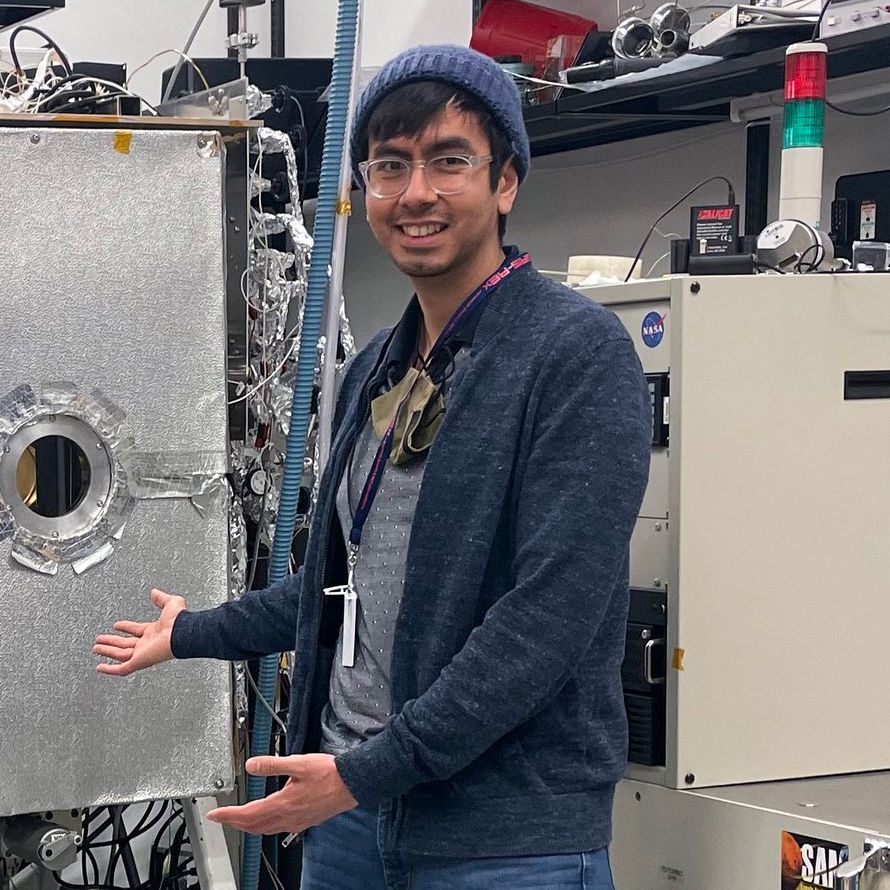 Man with glasses and a beanie smiling and gesturing towards equipment